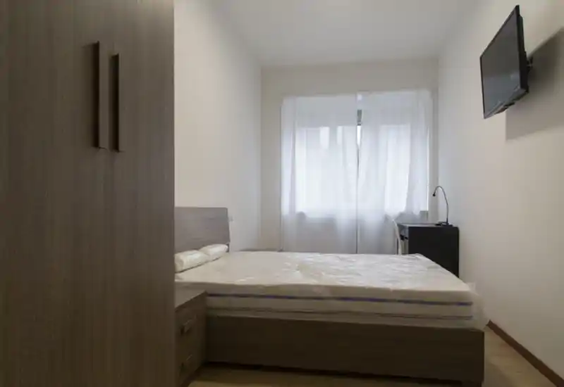 Chambre lumineuse et minimaliste avec lit double, table de chevet et télévision murale ; la lumière naturelle passe à travers des voilages.