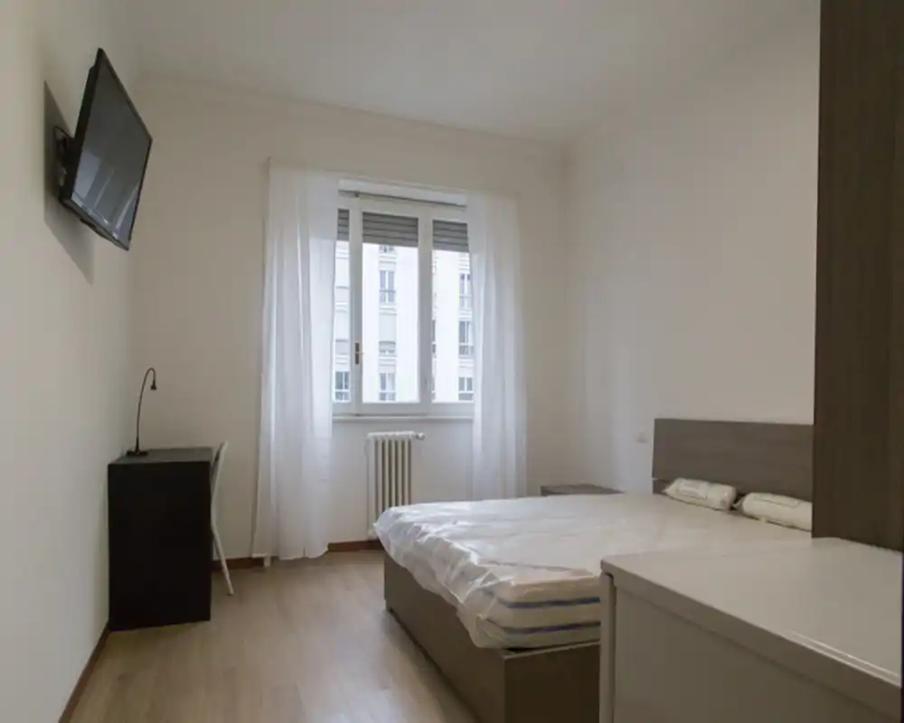 Camera da letto luminosa e minimale con letto matrimoniale, comodini e finestra con tende velate che lasciano entrare luce naturale. Scrivania semplice e TV a parete completano l’arredamento funzionale.