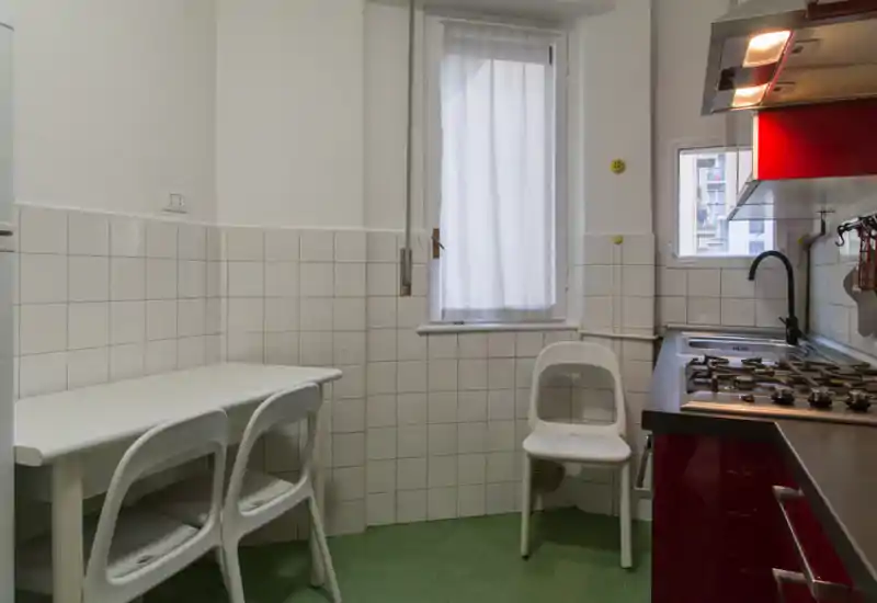 Cuisine compacte avec plaque à gaz, évier inox et petite table pour deux. La fenêtre apporte de la lumière naturelle et les meubles rouges ajoutent une touche moderne.