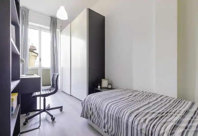 Camera singola moderna e luminosa con letto rifinito, ampia armadiatura, scrivania e luce naturale dalla finestra/terrazzino — ideale per studenti o professionisti.