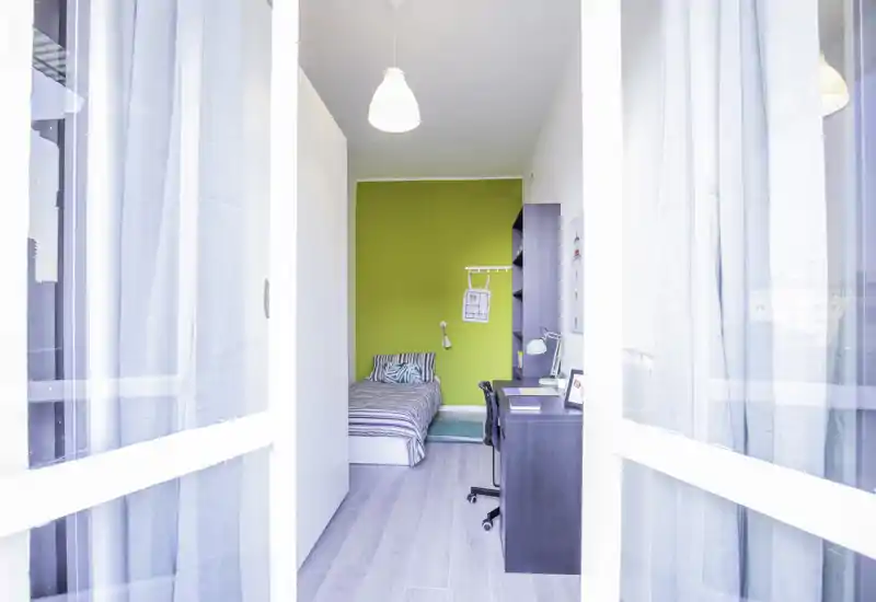 Camera singola luminosa con letto a righe, scrivania per lo studio e arredo moderno; grandi finestre garantiscono molta luce naturale.