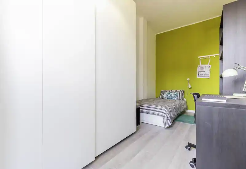 Camera singola compatta e luminosa con letto a righe, parete d'accento gialla, scrivania e grande armadio — ambiente ordinato e ideale per uno studente o una persona sola.