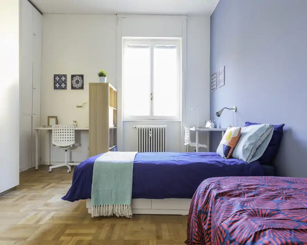Dormitorio luminoso y moderno con dos camas individuales, luz natural desde la ventana central, suelo de parquet y escritorios sencillos — ideal para alquiler compartido o estudiantes.