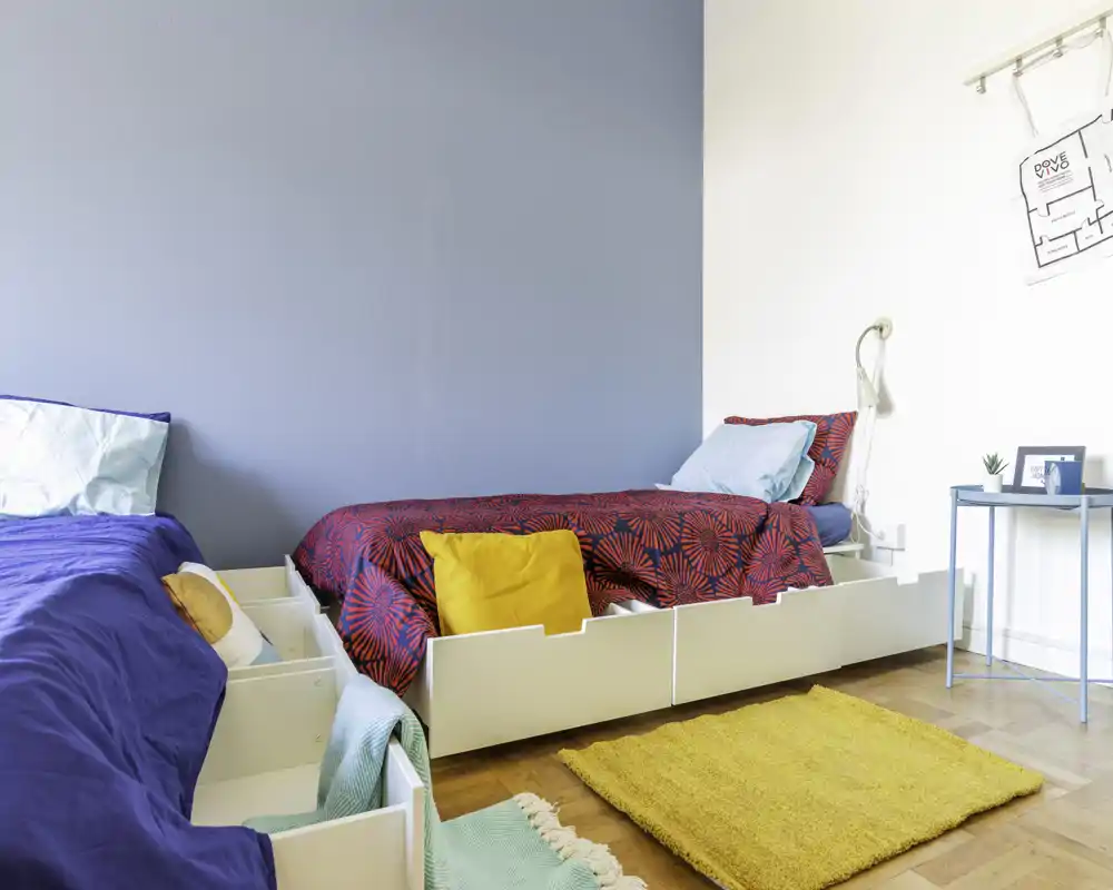 Dormitorio luminoso y colorido con dos camas individuales, ropa de cama vibrante y cojines; suelo de madera y mesita auxiliar que aportan un ambiente moderno y acogedor.