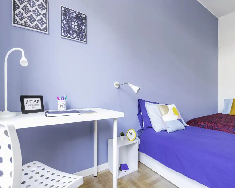 Dormitorio moderno y luminoso con dos camas individuales bien hechas frente a una pared azul, un pequeño escritorio blanco y detalles decorativos sencillos para una sensación fresca y acogedora.
