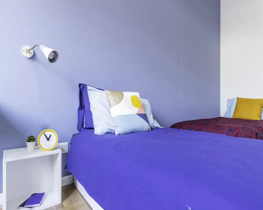 Dormitorio luminoso y moderno con una cama individual bien visible con ropa de cama azul, cojines decorativos y una mesita con reloj — ideal para alquileres de corta estancia.