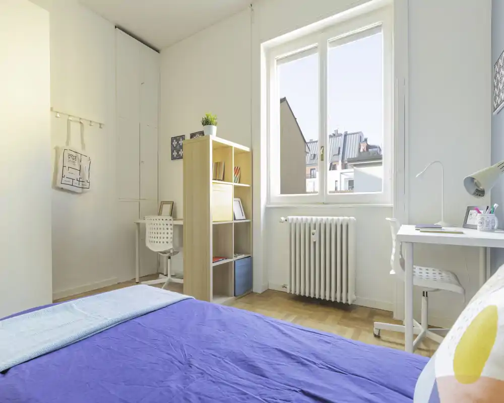 Dormitorio luminoso y ordenado con colcha azul, estantería sencilla y escritorio junto a la ventana — ideal para un estudiante o una persona sola.