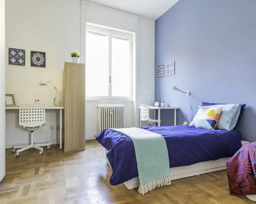 Dormitorio individual moderno y luminoso con la cama hecha, pared azul de acento y dos pequeños escritorios junto a la ventana — ideal para estudiante o una persona sola.