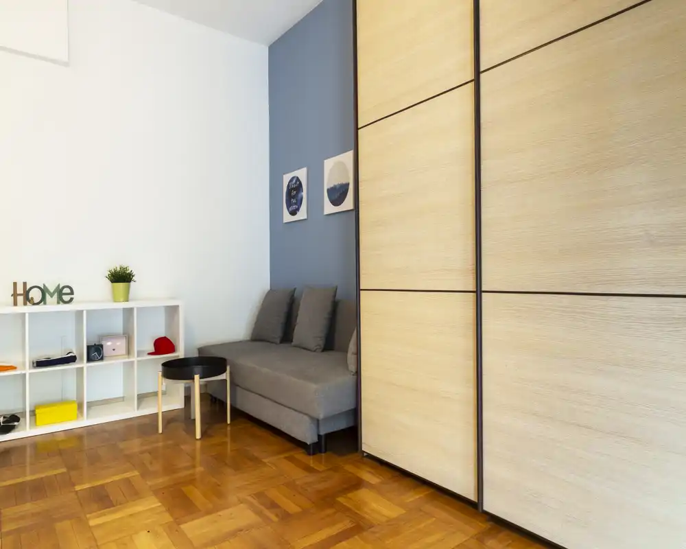 Área de estar moderna y ordenada con un sofá gris compacto, estantería decorativa y suelo de parquet cálido; luminosa y perfecta para un anuncio de apartamento pequeño.