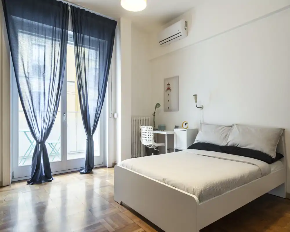 Dormitorio moderno y luminoso con cama doble, escritorio y grandes puertas de cristal que dan al balcón. Estilo minimalista, suelos de madera y luz natural suave que crean un ambiente tranquilo para alquileres.