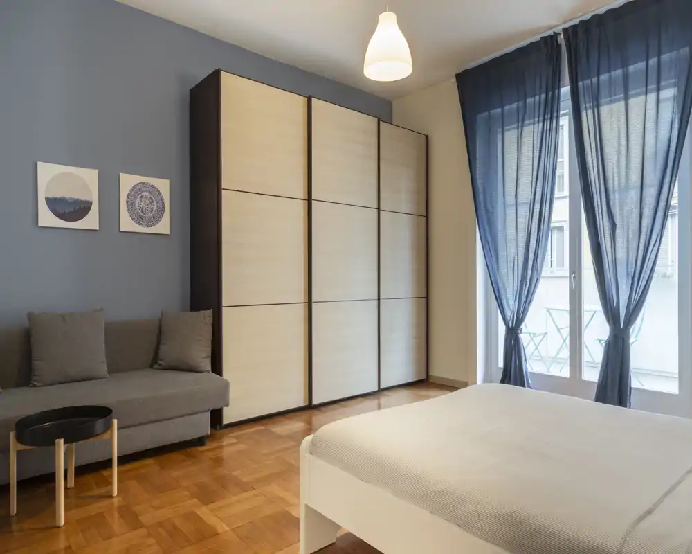 Dormitorio moderno y luminoso con cama doble, gran armario, sofá y acceso al balcón. Suelo de madera clara y textiles suaves que crean un ambiente sereno y acogedor.