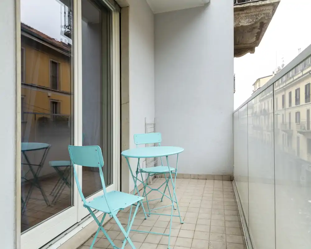 Pequeño balcón urbano con mesa tipo bistró turquesa y dos sillas a juego, suelo de baldosas y barandilla de cristal con vistas a edificios vecinos — rincón exterior luminoso y ordenado.