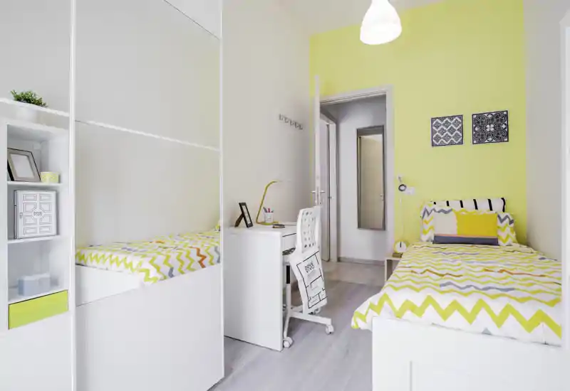Dormitorio individual moderno y luminoso con ropa de cama en zigzag, escritorio compacto y armario con puertas espejo — ambiente acogedor y bien decorado.