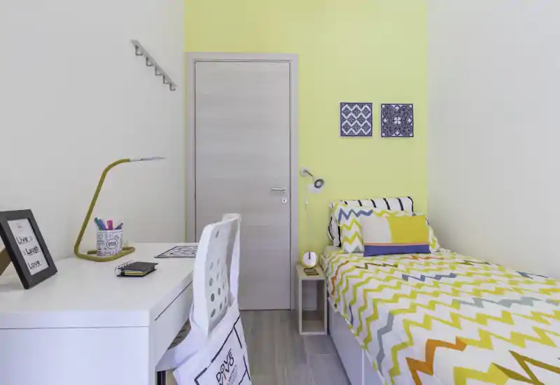 Dormitorio compacto y luminoso con cama individual, edredón de zigzag colorido, mesita de noche y escritorio ordenado. Las líneas limpias y la pared amarilla aportan un ambiente alegre, ideal para estudiantes o alquileres temporales.