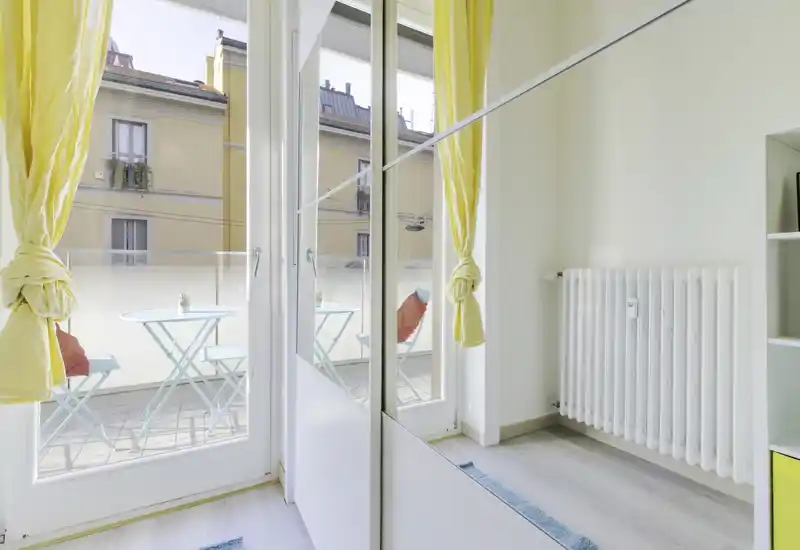 Pequeña terraza soleada con una mesa bistró y dos sillas, vista a través de grandes puertas de cristal con alegres cortinas amarillas — espacio exterior luminoso ideal para el café de la mañana.