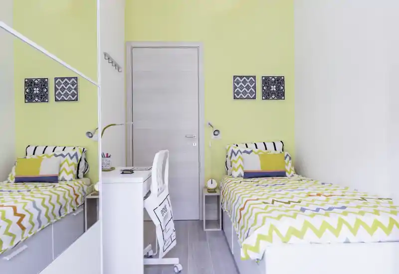 Dormitorio luminoso y moderno con dos camas individuales, ropa de cama con motivo zigzag y un pequeño escritorio entre ellas. El armario con espejo y los acentos amarillos coordinados aportan una sensación fresca y aireada, ideal para alquileres temporales.