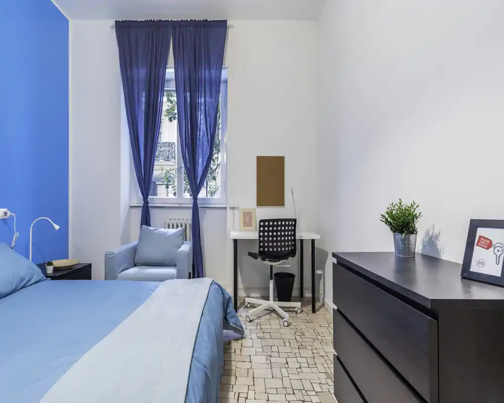 Bright, well-staged bedroom with a blue theme: comfortable bed, matching armchair, desk and dresser. Natural light from the window and simple modern decor make it ideal for short-term rentals.