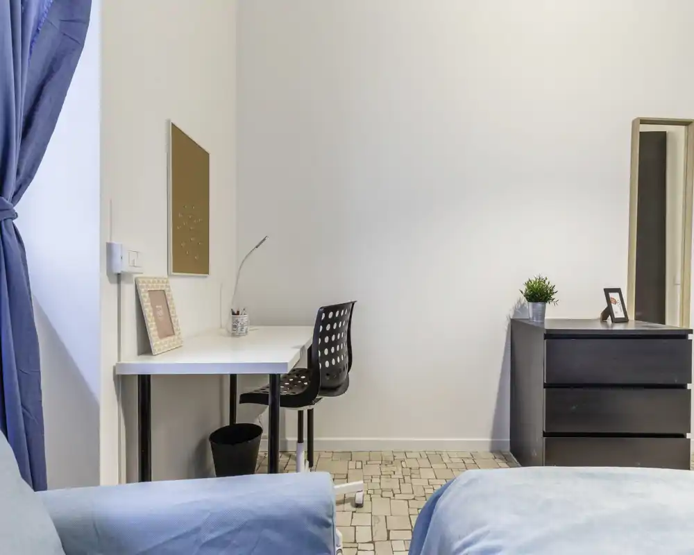 Bright, simply furnished bedroom corner with two beds partially visible, a compact desk and chair, and a small chest of drawers with a plant — suitable for a cozy single or shared room.