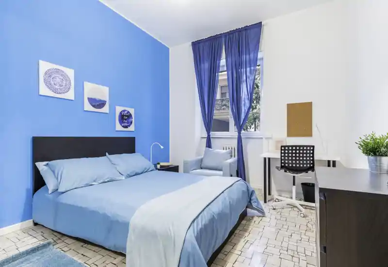 Bright, modern bedroom with a double bed dressed in blue linens, coordinated curtains and a cozy armchair by the window. The room features a desk area suitable for work and tasteful wall art, offering a fresh, well-styled space.