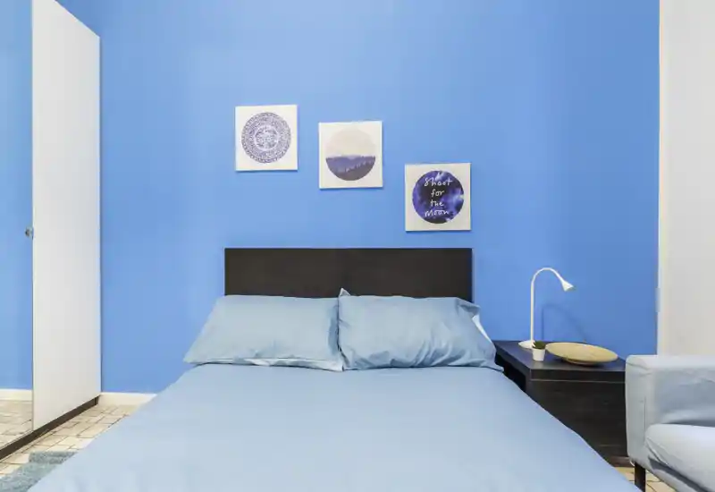 Bright, modern bedroom with a full bed dressed in blue linens, coordinated wall art and a bedside table with lamp — vivid, well-styled space ideal for listings.