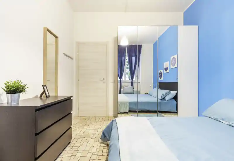 Bright, modern bedroom with a blue accent wall, double bed dressed in matching linens, mirrored wardrobe and a dark chest of drawers; airy and well-lit space ideal for listings.
