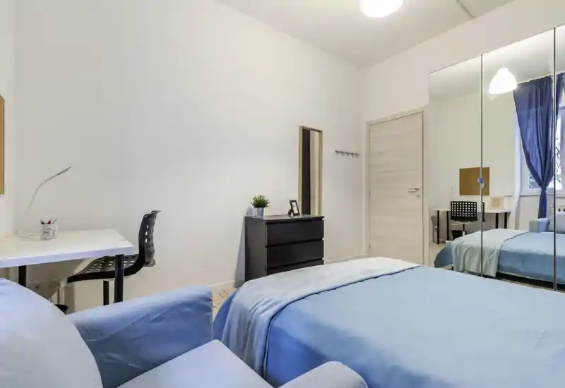 Bright, modern bedroom with a neatly made double bed in blue linens, a desk and chair for study, and mirrored sliding wardrobes that add depth and light.