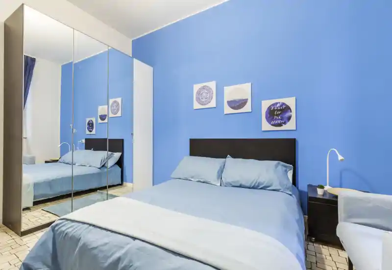 Bright, modern bedroom with a double bed dressed in light-blue linens, a dark headboard, mirrored wardrobe and simple wall art on a vivid blue wall.