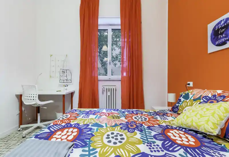 Colorful bedroom with a large patterned bed, red curtains and a small workspace by the window. Bright, well-lit room with lively textiles and modern accents.
