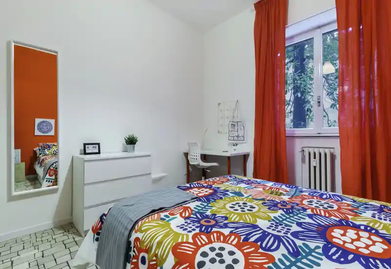 Colorful, well-lit bedroom with a patterned bedspread, a small desk by the window, dresser and full-length mirror. Bright red curtains frame the window and the room feels fresh and welcoming — ideal for a single or student rental.