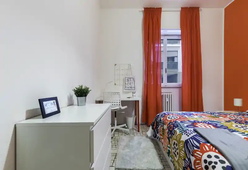 Bright, colorful bedroom with a patterned bedspread, bedside lamp and a small workspace by the window with red curtains that add character.
