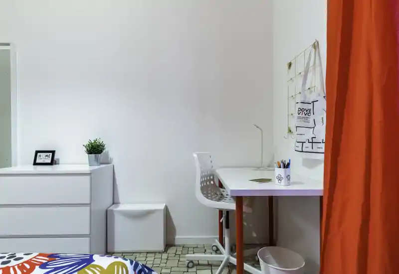 Bright, modern bedroom corner with a white desk and chair, chest of drawers, and a colorful bedspread. Clean styling, useful workspace and cheerful red curtain give a fresh, inviting feel.