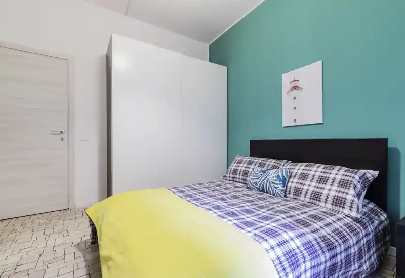 Bright, modern bedroom with a double bed framed by a dark headboard and a vibrant turquoise accent wall; plaid bedding, decorative cushion and a white wardrobe complete the clean, welcoming space.