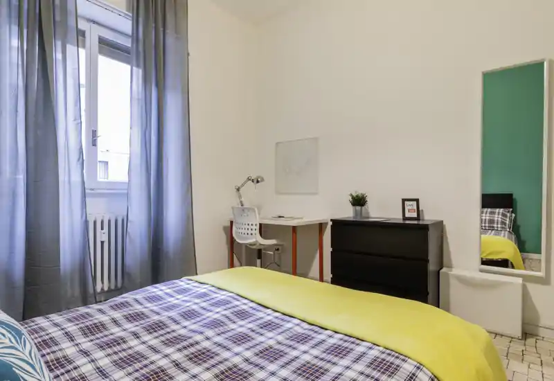 Bright, neatly styled bedroom with a patterned bedspread, yellow throw and a study corner by the window. The room features a tall mirror, chest of drawers and soft natural light.