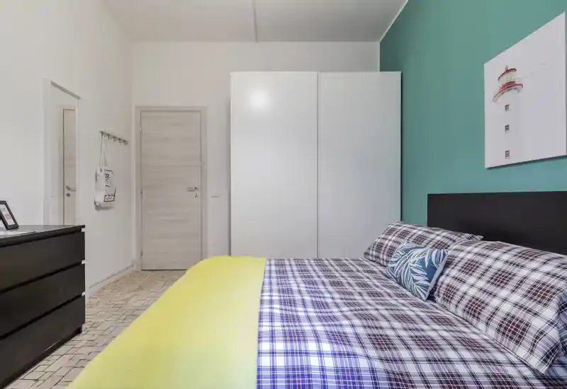 Bright, neatly staged bedroom with a large bed dressed in checked linens and a yellow throw, a white wardrobe and teal accent wall creating a fresh, modern look.