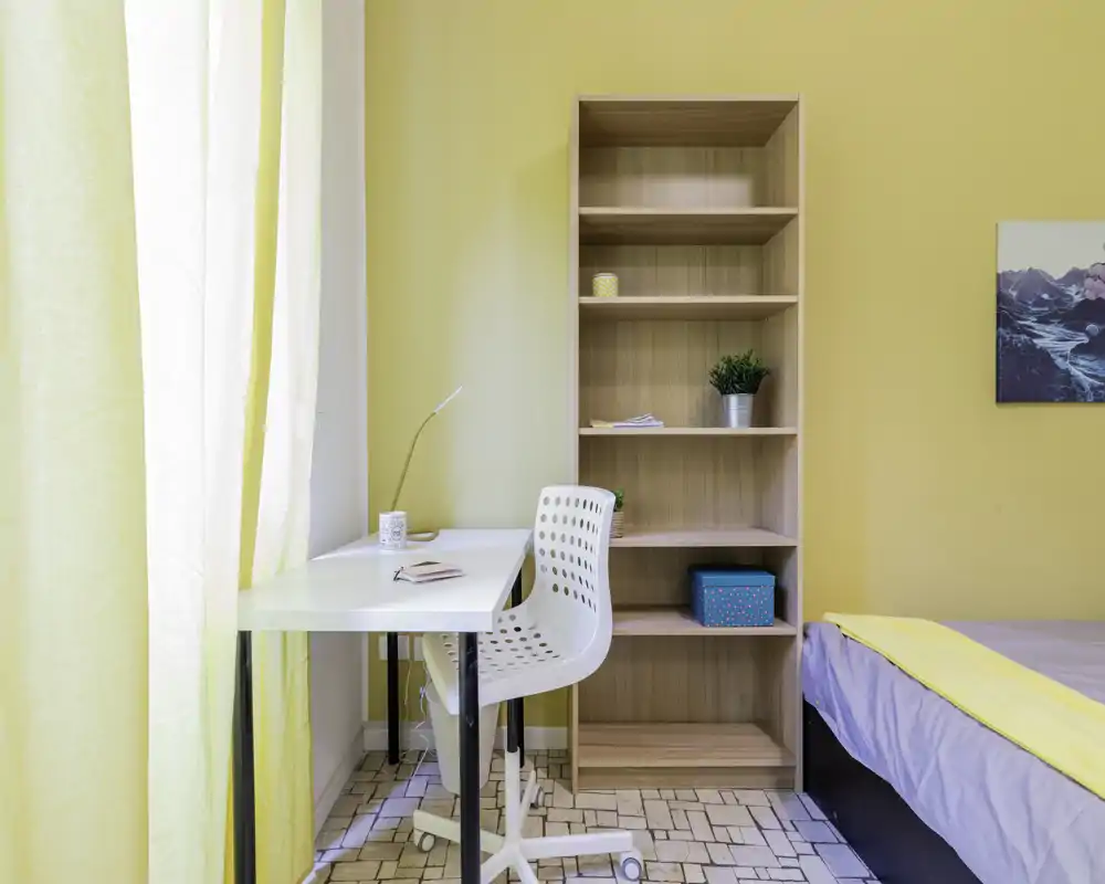 Dormitorio pequeño y soleado con cama individual, escritorio junto a la ventana y estantería abierta; paredes amarillas y luz natural que aportan calidez.