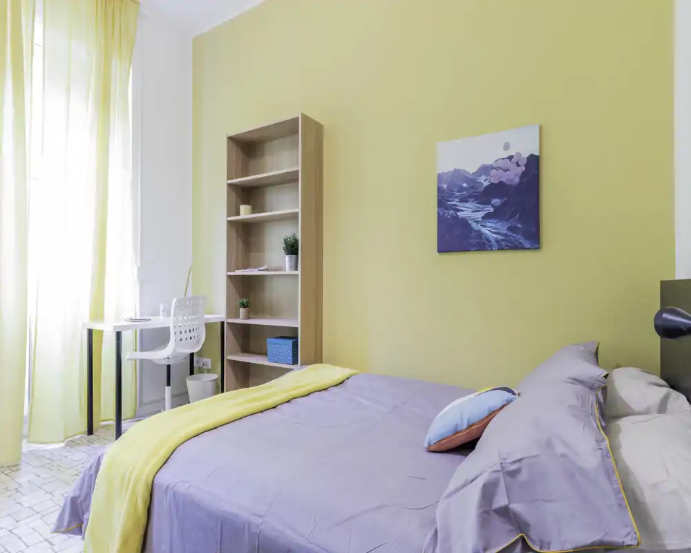 Dormitorio moderno y luminoso con cama doble bien hecha, pequeño escritorio junto a la ventana, estantería abierta y pared amarilla de acento—ideal para una estancia cómoda con buena luz natural.