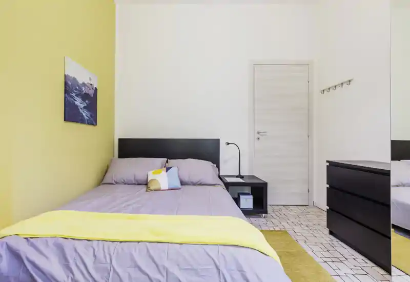 Camera da letto luminosa e curata con letto matrimoniale, comodino e comò coordinati; parete d'accento gialla e biancheria morbida che creano un'atmosfera accogliente.