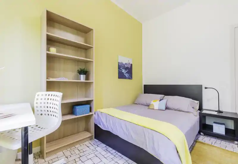 Camera da letto luminosa e accogliente con letto matrimoniale, comodino e scaffalatura; parete gialla e tessili curati creano un'atmosfera invitante.