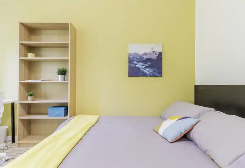 Camera da letto luminosa e curata con letto ben rifatto, cuscino e coperta colorati e una libreria a vista su una parete gialla calda.