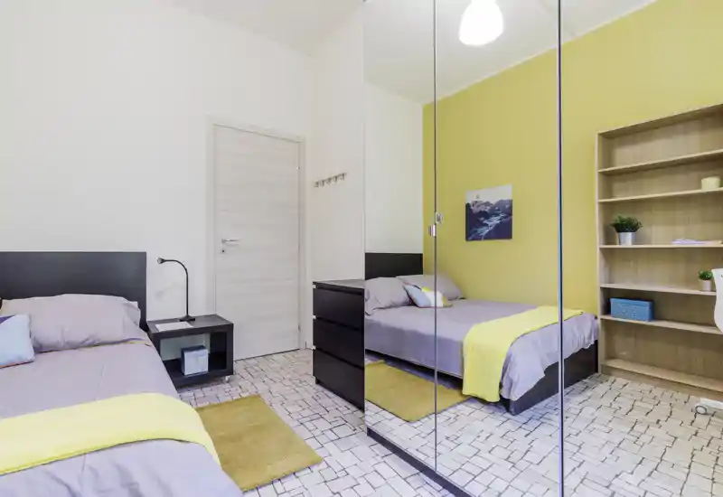 Camera da letto moderna e luminosa con letti singoli, armadio con ante a specchio che amplia lo spazio, tessili accoglienti e parete d'accento gialla.