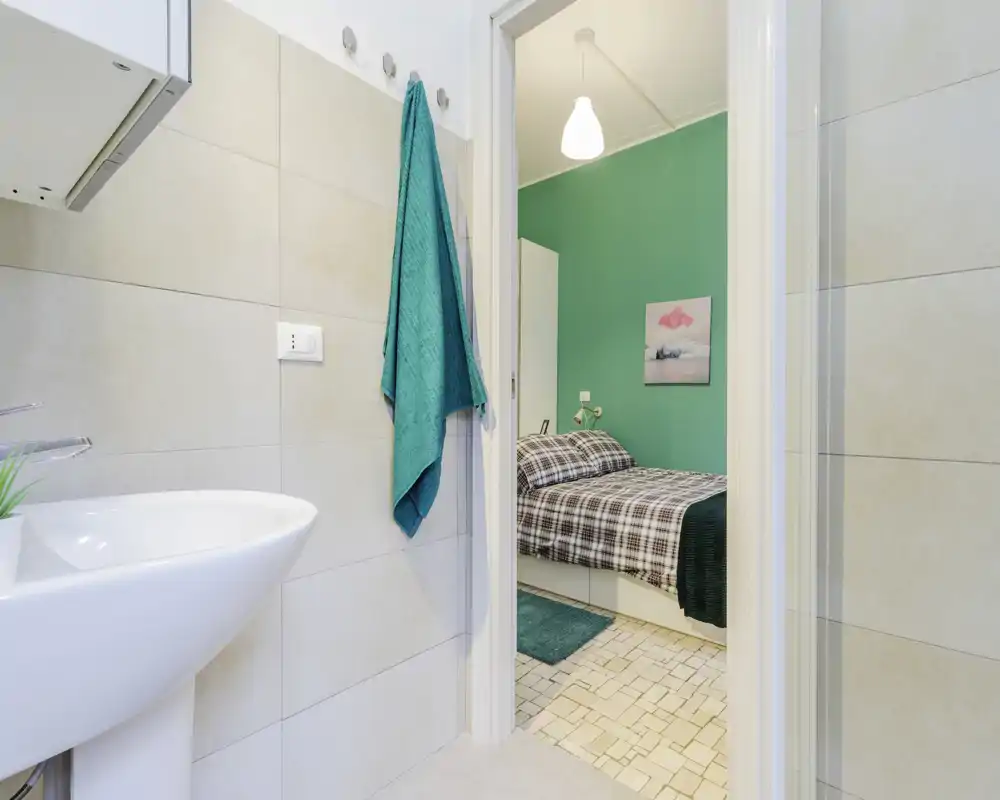 Vue depuis la salle de bain vers une petite chambre : un lit double avec couette à carreaux et des accents turquoise visible par la porte, agencement pratique et coordonné.