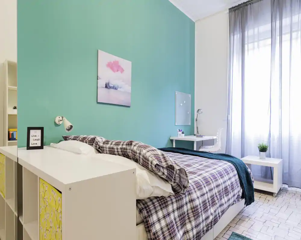 Chambre lumineuse et stylée avec un mur d'accent turquoise, grand lit avec couette à motifs, lampe de chevet et un petit bureau près d'une grande fenêtre offrant beaucoup de lumière naturelle.
