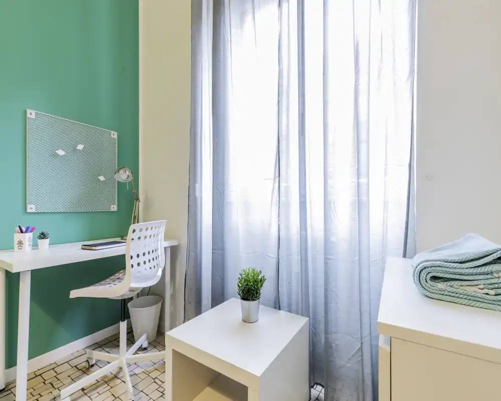 Coin chambre lumineux et minimaliste avec bureau et petite table : lumière naturelle à travers des voilages, espace de travail compact et touches décoratives soignées.