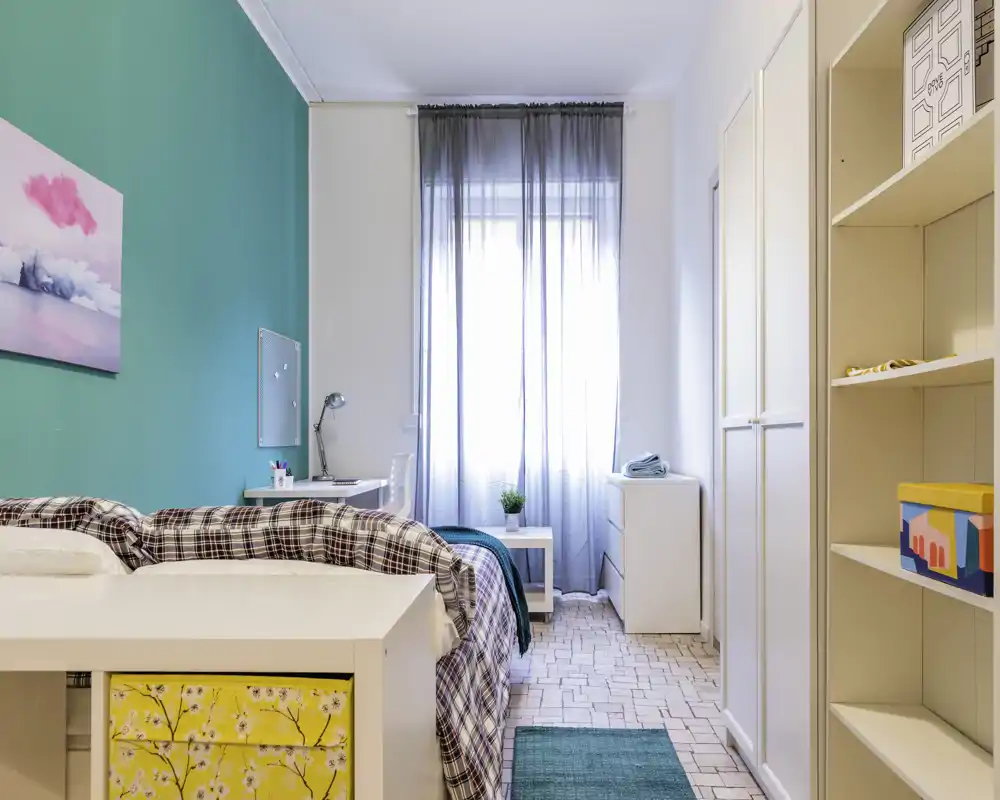 Chambre lumineuse et bien aménagée avec lit complet, bureau et armoire ; un mur accent turquoise et la lumière naturelle apportent une ambiance fraîche et accueillante.