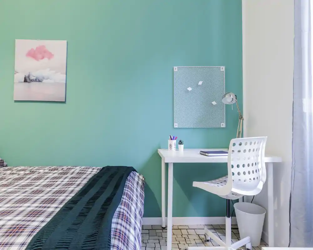 Chambre moderne et lumineuse avec un lit bien fait et un petit bureau contre un mur accent turquoise. Lignes épurées, lumière naturelle et textiles assortis offrent un espace accueillant, idéal pour la location courte durée.