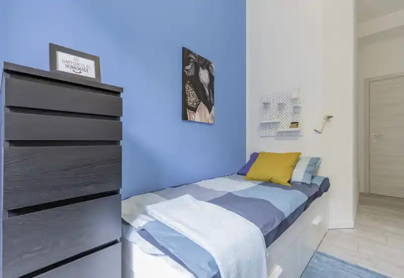 Bright, modern single bedroom with a neatly made bed against a vivid blue accent wall, decorative cushions, wall art and a tall chest of drawers—ideal for a compact rental.