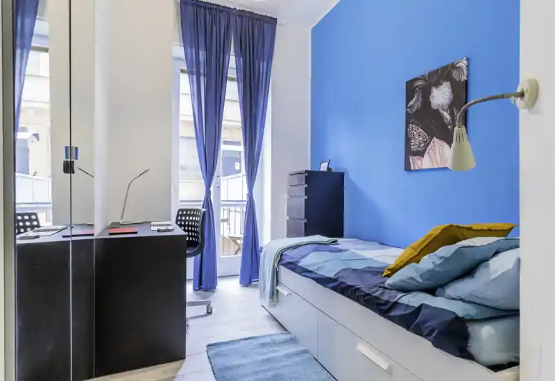 Bright single bedroom with a turquoise accent wall, comfortable bed with layered bedding, small desk and tall windows letting in ample natural light — ideal for a student or single occupant.