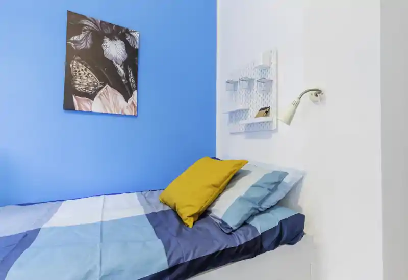 Small, bright bedroom corner with a neatly made single bed, colorful cushions and modern wall lamp — ideal for a cozy solo stay.
