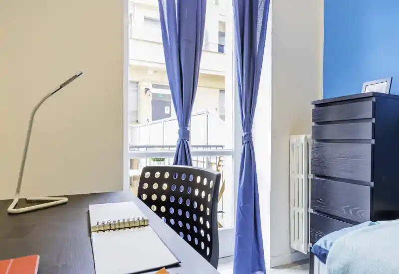 Bright, modern bedroom corner with a study desk, blue curtains and a chest of drawers; natural light floods the room through a large window.