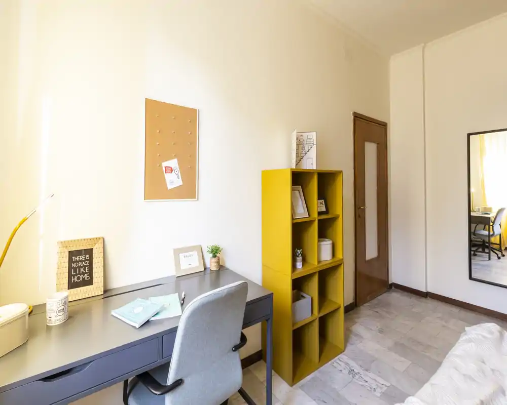 Angolo camera luminoso e ordinato con scrivania e sedia accanto a un mobile giallo a cubi; parte del letto è visibile sul bordo destro. Lo spazio è ben illuminato e arredato con semplicità, ideale per studiare o lavorare da casa.